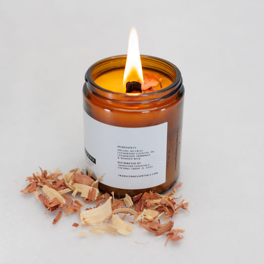 A lit amber glass candle sits on a light surface with cedarwood shavings scattered in front of it. The label lists ingredients including organic beeswax, cedarwood essential oil, cedarwood trimmings, and a wooden wick, along with distributor details for Transcend Essentials.
