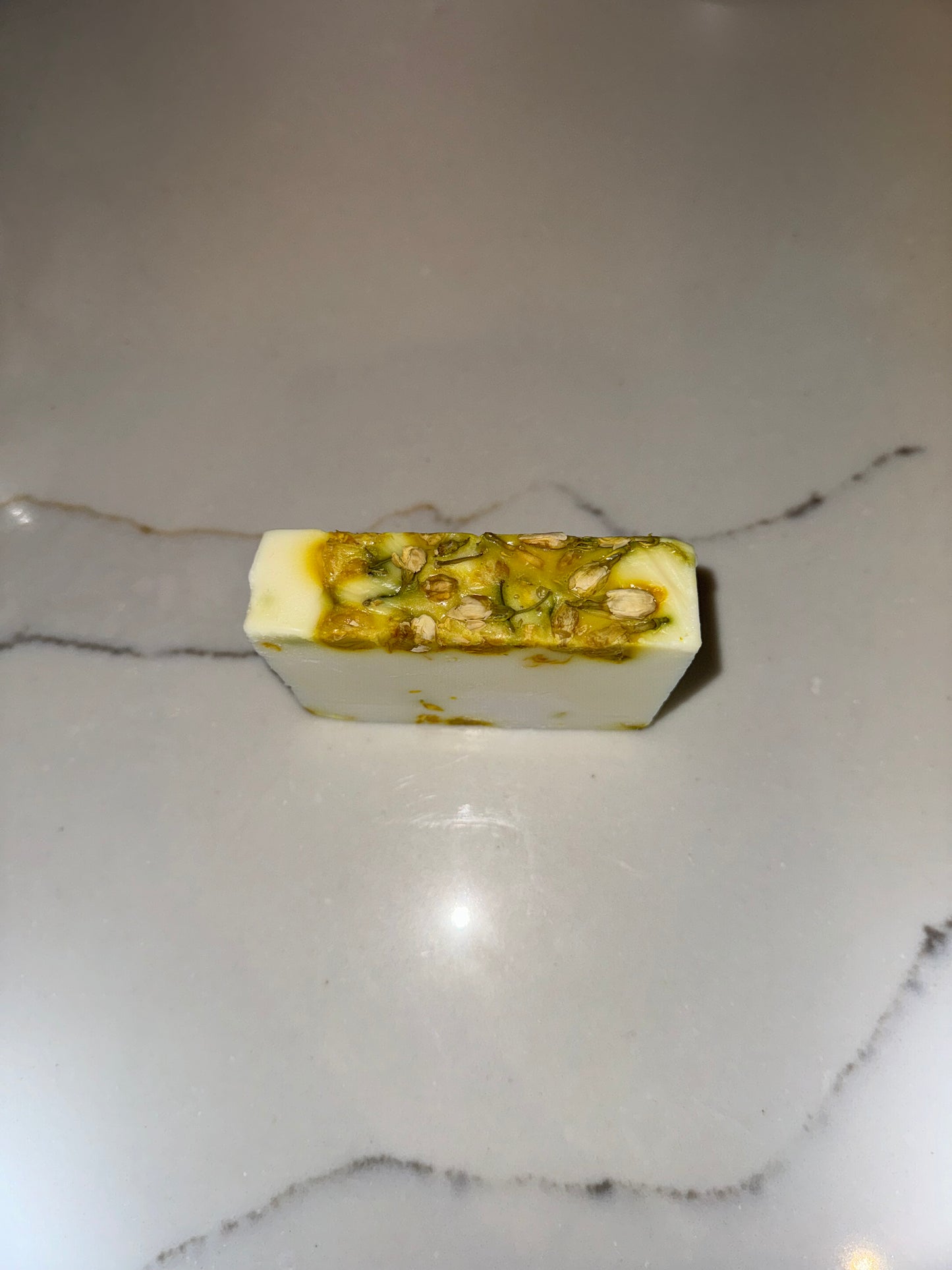 The image shows a bar of Transcend Hydrating & Soothing Jasmine Bar Soap. The soap is predominantly white with green and brown flecks, indicating the presence of jasmine and other natural ingredients. It is placed on a light-colored countertop with a subtle vein pattern. The soap's appearance highlights its hydrating and soothing properties, suggesting a calming and natural bathing experience.