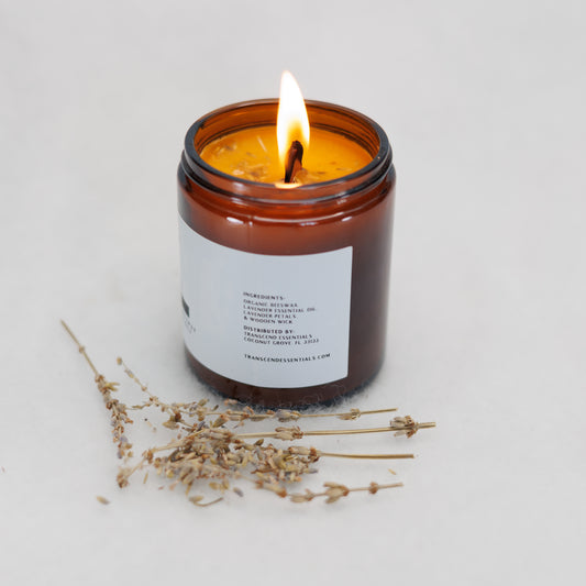 A lit amber glass candle showing the ingredients label sits on a light surface, with dried lavender sprigs placed in front of it. The label lists the ingredients as organic beeswax, lavender essential oil, lavender petals, and a wooden wick. The candle is made by Transcend Essentials.