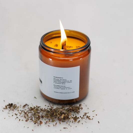 A lit amber glass candle sits on a light surface with dried peppermint leaves scattered in front of it. The label lists ingredients including organic beeswax, peppermint essential oil, dried peppermint leaves, and a wooden wick, along with distributor details for Transcend Essentials.