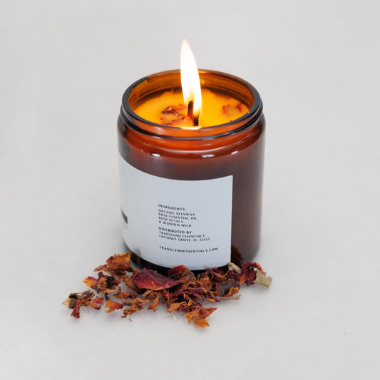 A lit amber glass candle sits on a light surface with dried rose petals scattered in front of it. The label lists ingredients including organic beeswax, rose essential oil, dried rose petals, and a wooden wick, along with distributor details for Transcend Essentials.