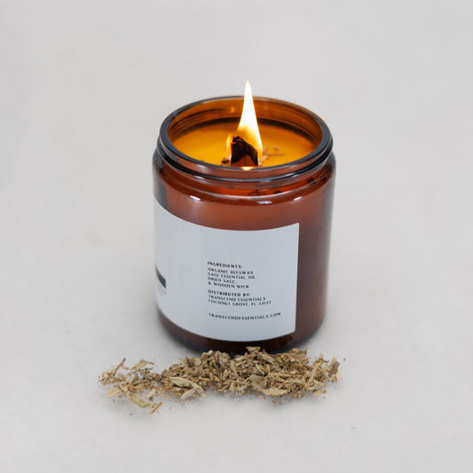 A lit amber glass candle sits on a light surface with a small pile of dried sage pieces in front of it. The candle’s label shows the ingredients, including organic beeswax, sage essential oil, dried sage, and a wooden wick, along with the distributor’s information for Transcend Essentials.
