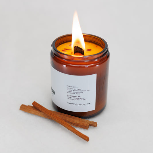 A lit amber glass candle sits on a light surface with two pieces of sandalwood placed in front of it. The label lists ingredients including organic beeswax, sandalwood essential oil, sandalwood chips, and a wooden wick, along with distributor details for Transcend Essentials.