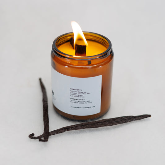 A lit amber glass candle showing the ingredients label sits on a light surface, with two vanilla beans placed in front of it. The label lists the ingredients as organic beeswax, vanilla essential oil, vanilla bean, and a wooden wick. The candle is made by Transcend Essentials.