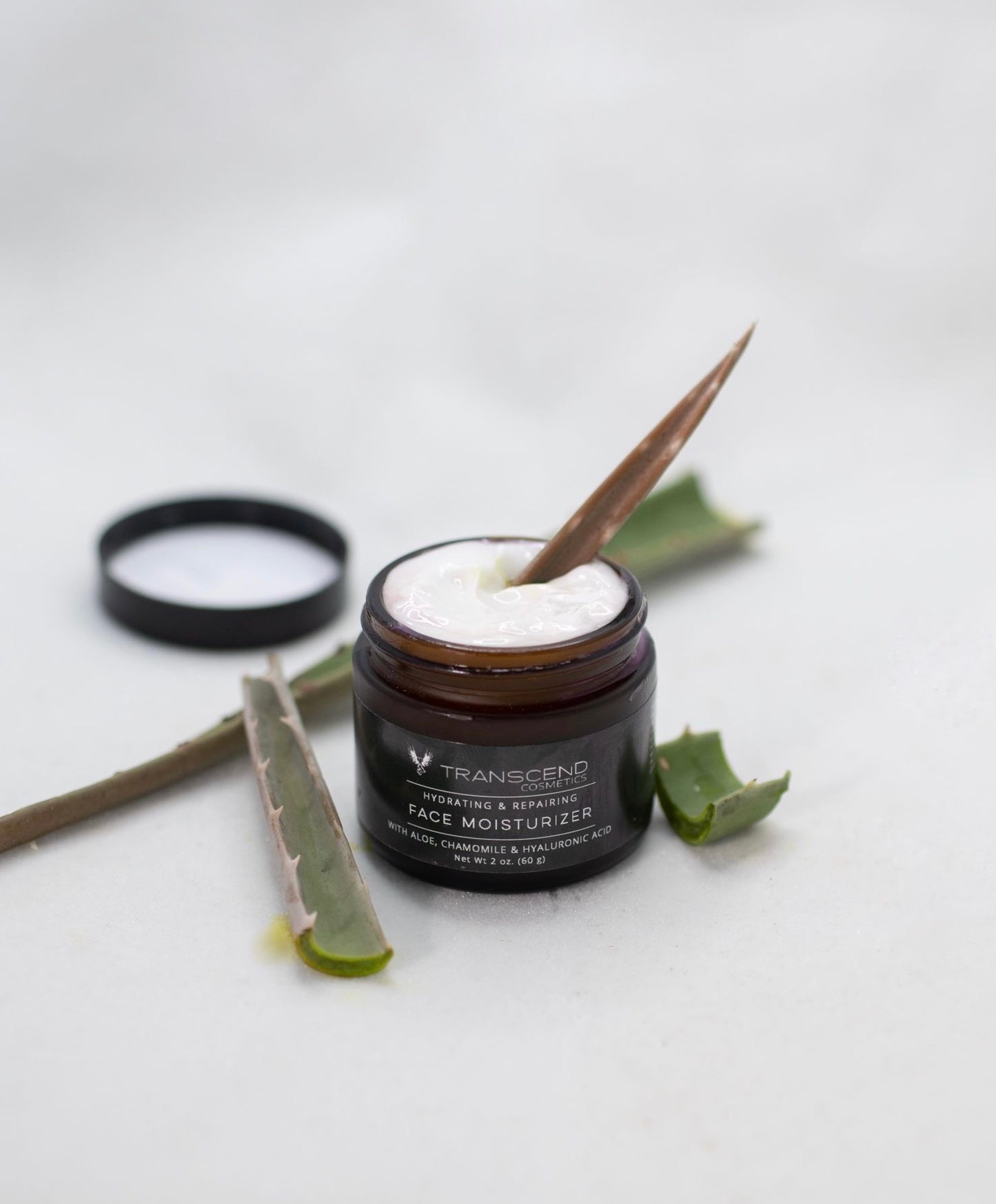 The image features a jar of Transcend Hydrating & Repairing Face Moisturizer on a light background. The moisturizer is creamy and white, with a aloe Vera leaf inside resting in the cream, lifting some of the product. Beside the jar, there are a few fresh aloe vera leaves, one of which is sliced open to show the gel inside. The product label indicates it contains aloe, chamomile, and hyaluronic acid.