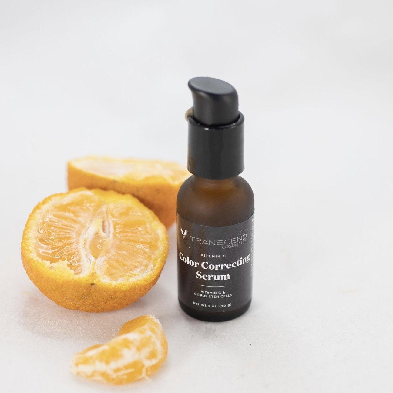The image shows a small dark amber bottle of Transcend Vitamin C Color Correcting Serum on a white background, accompanied by a halved orange and a segment of orange. The serum bottle has a black pump dispenser and is labeled clearly, emphasizing its key ingredients, Vitamin C and citrus stem cells. The fresh orange beside it highlights the natural citrus elements used in the serum.Natural Skincare made with Oranges.