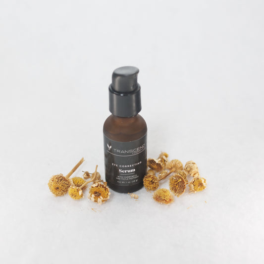 The image shows a dark amber bottle of Transcend Eye Correcting Serum, surrounded by dried chamomile flowers on a white background. The serum bottle features a black pump dispenser, and the label on the bottle indicates it contains chamomile and triple peptides. The flowers are scattered around the base of the bottle, emphasizing the natural ingredients theme.  Natural skincare made with chamomile. 
