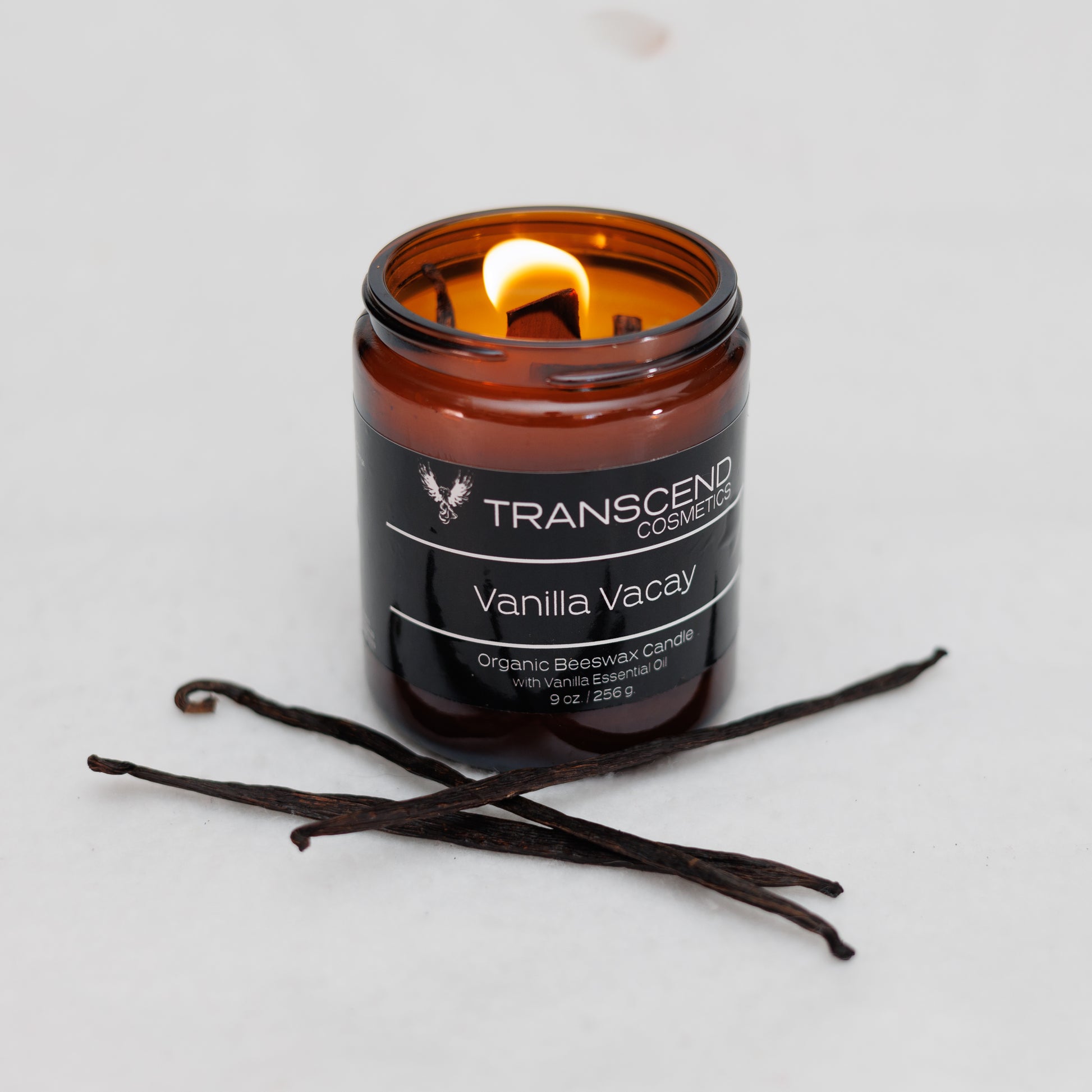 Transcend Cosmetics Vanilla Vacay Organic Beeswax Candle in an amber glass jar with a wooden wick, burning with a warm flame, surrounded by vanilla beans, highlighting its natural and soothing ingredients.