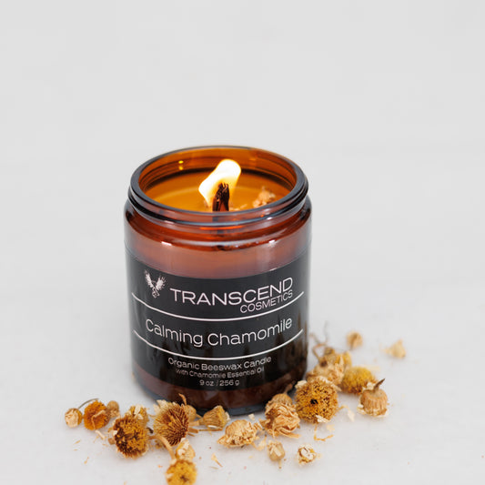 Transcend Cosmetics Calming Chamomile Organic Beeswax Candle in an amber glass jar, with a wooden wick and dried chamomile flowers scattered around, creating a soothing and natural ambiance.