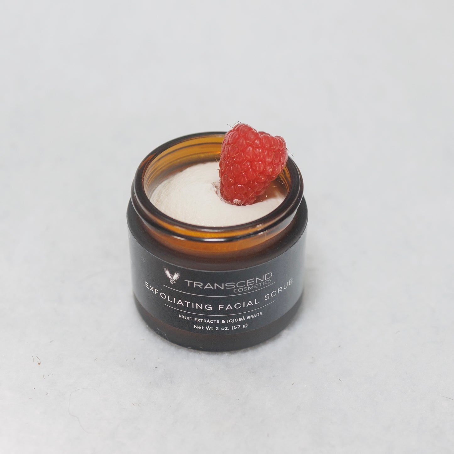 The image shows a jar of Transcend Exfoliating Facial Scrub on a light background. The scrub appears creamy with a fresh raspberry placed on top. The jar is dark brown with a label stating it contains fruit extracts and jojoba beads. This product presentation highlights the natural ingredients used in the scrub.
