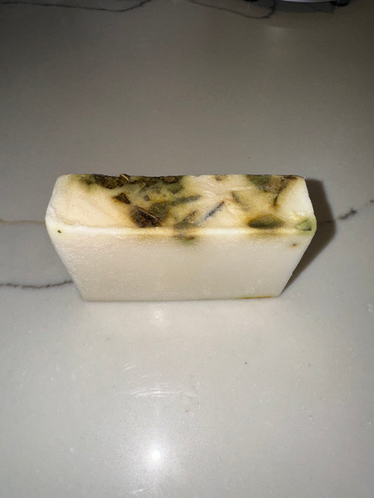 The image shows a bar of Transcend Hydrating & Invigorating Eucalyptus Bar Soap. The soap is white with visible green and brown flecks, indicating the presence of eucalyptus and other natural ingredients. It is placed on a light-colored countertop with a subtle vein pattern. The soap's appearance highlights its hydrating and invigorating properties, suggesting a refreshing and natural bathing experience