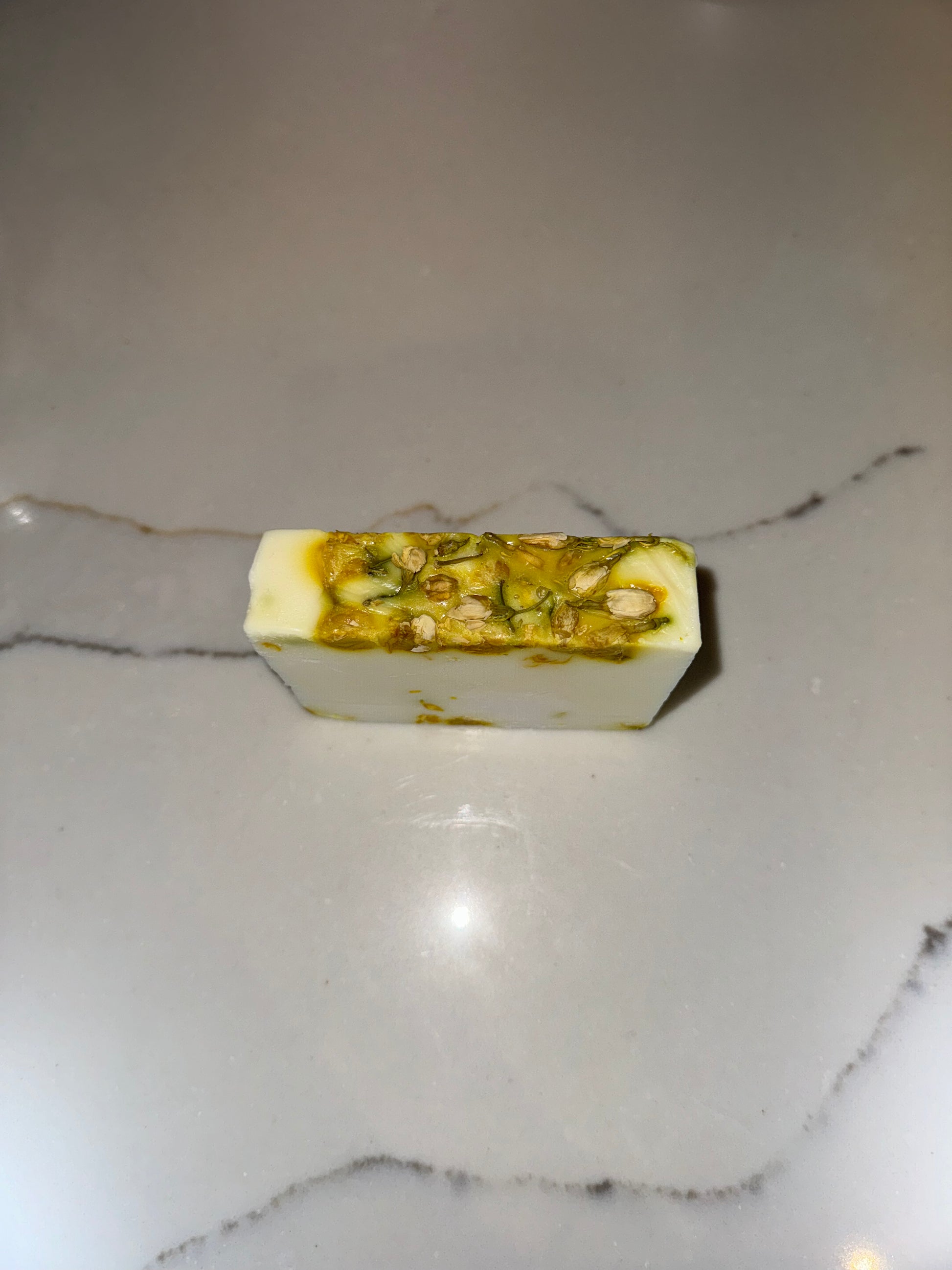 The image shows a bar of Transcend Hydrating & Soothing Jasmine Bar Soap. The soap is predominantly white with green and brown flecks, indicating the presence of jasmine and other natural ingredients. It is placed on a light-colored countertop with a subtle vein pattern. The soap's appearance highlights its hydrating and soothing properties, suggesting a calming and natural bathing experience.
