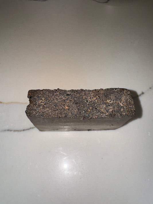 The image shows a bar of Transcend Hydrating & Exfoliating Coffee Bar Soap with a rough, textured surface. The soap is dark brown, suggesting the inclusion of coffee grounds for exfoliation. It is placed on a light-colored countertop with a subtle vein pattern. The rugged, earthy appearance highlights its natural and exfoliating properties.
