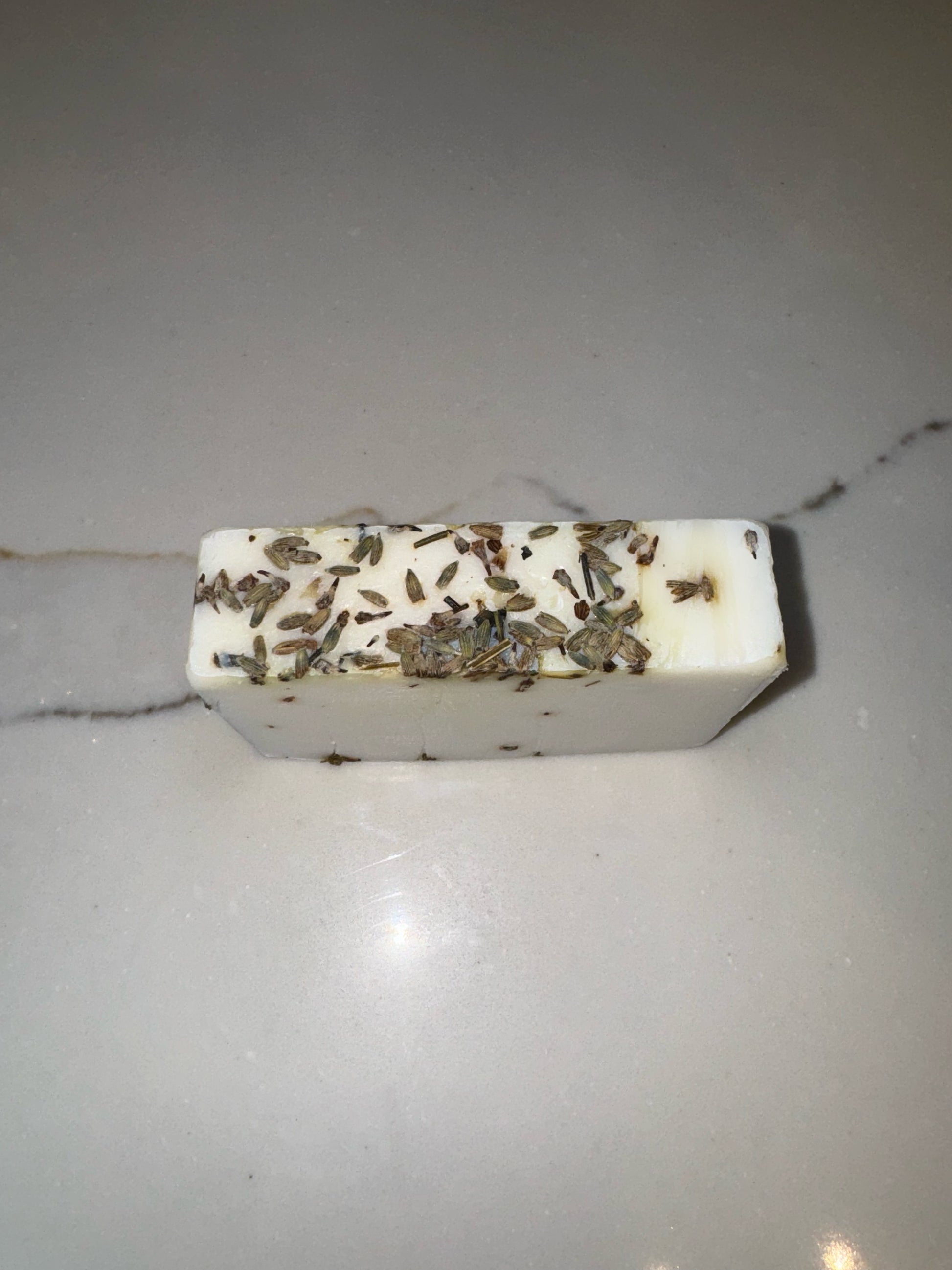 The image shows a bar of Transcend Hydrating & Calming Lavender Bar Soap with dried lavender buds embedded on its surface. The soap is placed on a light-colored countertop with a subtle vein pattern. The dried lavender adds a natural, decorative element to the soap, highlighting its botanical ingredients.
