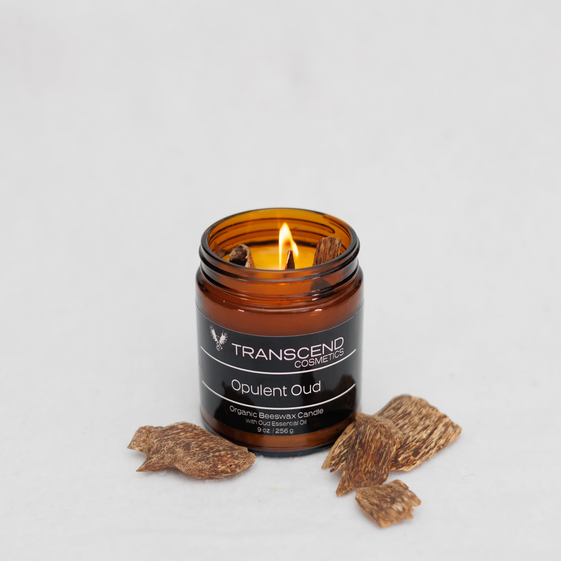 Transcend Cosmetics Opulent Oud Organic Beeswax Candle in an amber glass jar with a wooden wick, burning with a warm flame, surrounded by Oud Wood Chips, highlighting its natural and soothing ingredients.