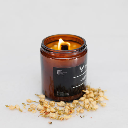 Image showing the ingredient list on the Transcend Cosmetics Jasmine Blossom Organic Beeswax Candle, in an amber glass jar with a wooden wick, burning with a warm flame, surrounded by dried Jasmine flowers, highlighting its natural and soothing ingredients.