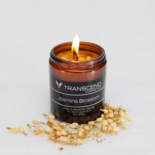 Transcend Cosmetics Soothing Sage Organic Beeswax Candle in an amber glass jar with a wooden wick, burning with a warm flame, surrounded by dried Jasmine flowers, highlighting its natural and soothing ingredients.