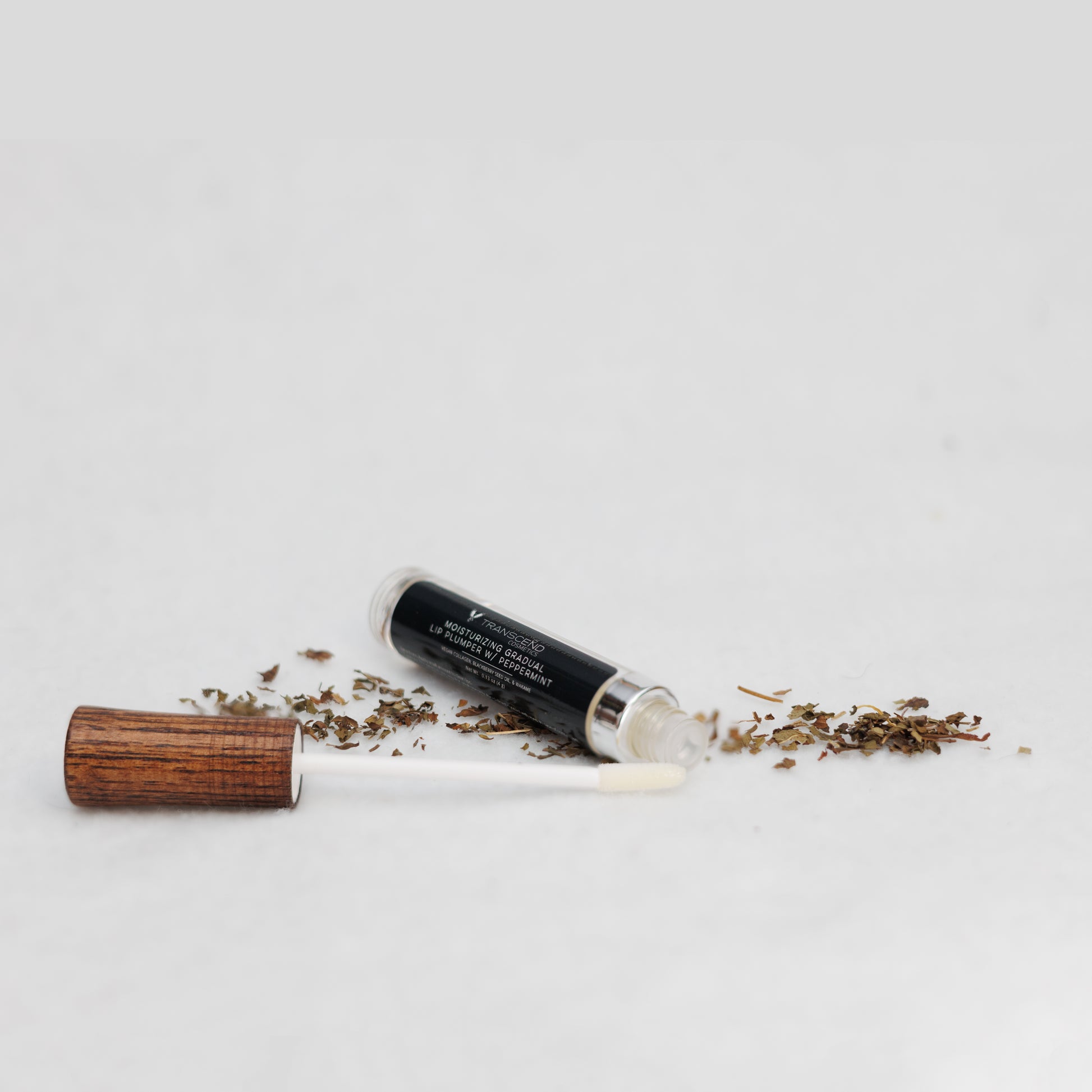 Transcend Cosmetics moisturizing gradual lip plumper with peppermint, featuring a sleek design with a wooden applicator handle, placed on a white surface with dried peppermint leaves scattered around.