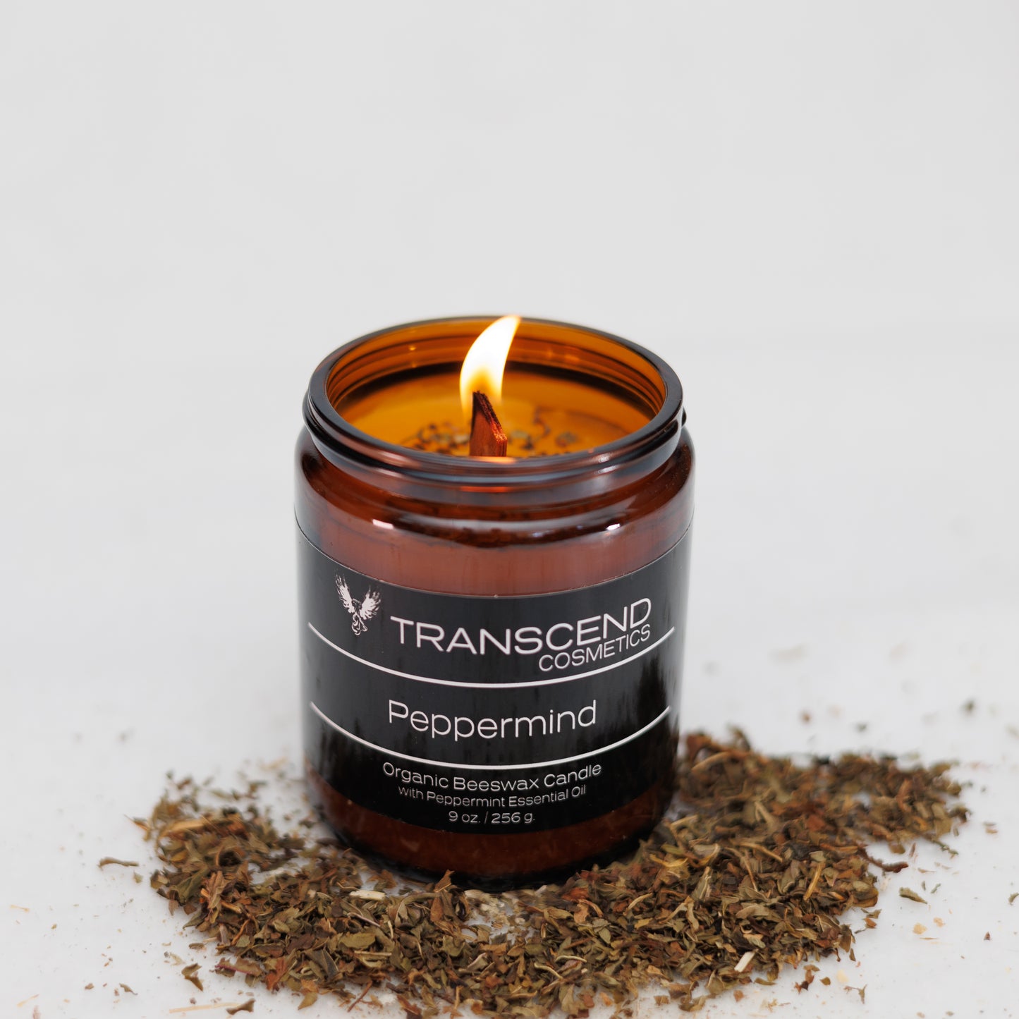 Transcend Cosmetics Peppermind Organic Beeswax Candle in an amber glass jar with a wooden wick, burning with a warm flame, surrounded by dried Peppermint leaves, highlighting its natural and soothing ingredients.