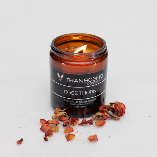 Transcend Cosmetics Rose Thorn Organic Beeswax Candle in an amber glass jar with a wooden wick, burning with a warm flame, surrounded by dried Rose petals, highlighting its natural and soothing ingredients.