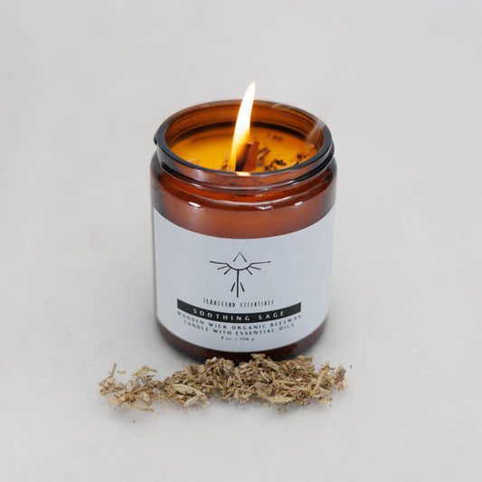 A lit amber glass candle labeled “Transcend Essentials – Soothing Sage” sits on a light surface, with a small pile of dried sage pieces in front of it. The candle has a wooden wick and is made from organic beeswax with essential oils.