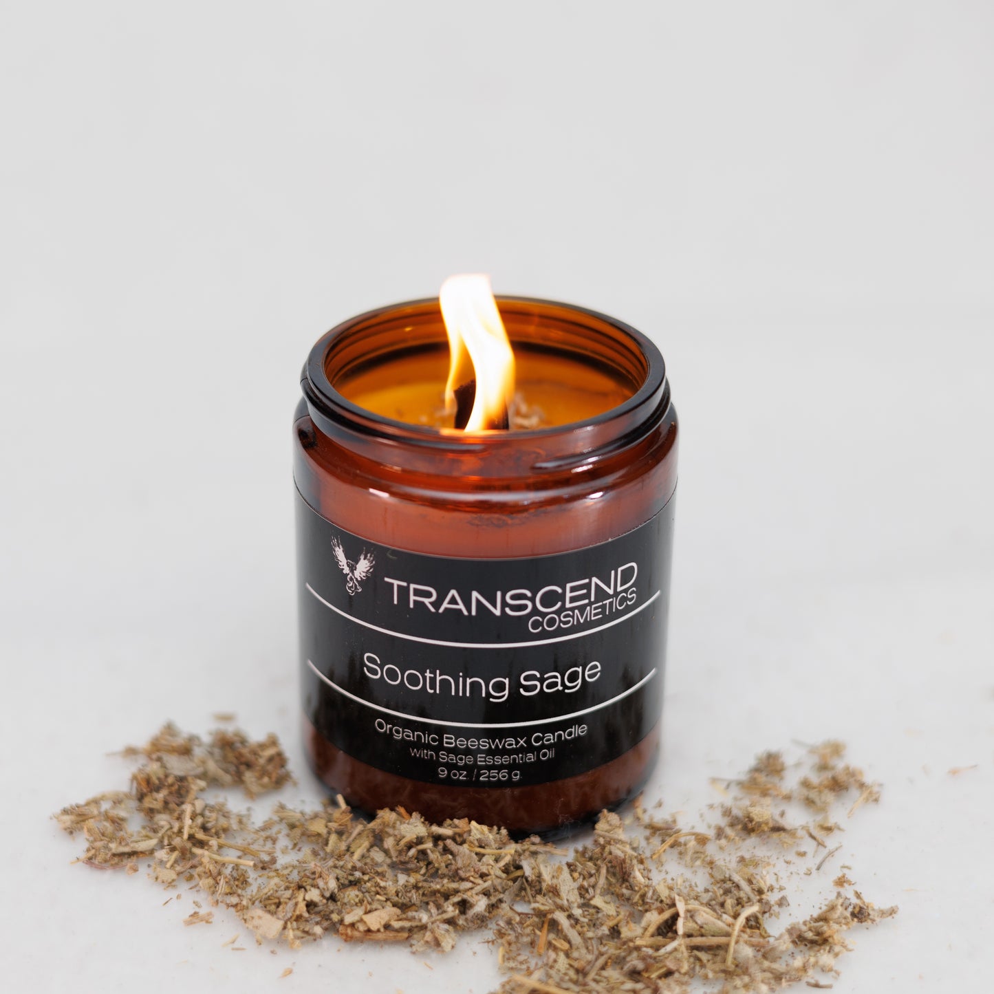 Transcend Cosmetics Soothing Sage Organic Beeswax Candle in an amber glass jar with a wooden wick, burning with a warm flame, surrounded by dried sage flowers, highlighting its natural and soothing ingredients.