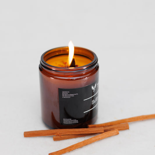 Image showing the ingredient list on the Transcend Cosmetics Sultry Sandalwood Organic Beeswax Candle, in an amber glass jar with a wooden wick, burning with a warm flame, surrounded by Sandalwood sticks, highlighting its natural and soothing ingredients.
Ingredients are: Beeswax, Sandalwood Essential Oil, Sandalwood Sticks, & Wooden Wick. 