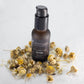 The image shows a dark amber bottle of Transcend Eye Correcting Serum, surrounded by dried chamomile flowers on a white background. The serum bottle features a black pump dispenser, and the label on the bottle indicates it contains chamomile and triple peptides. The flowers are scattered around the base of the bottle, emphasizing the natural ingredients theme.  Natural skincare made with chamomile. 
