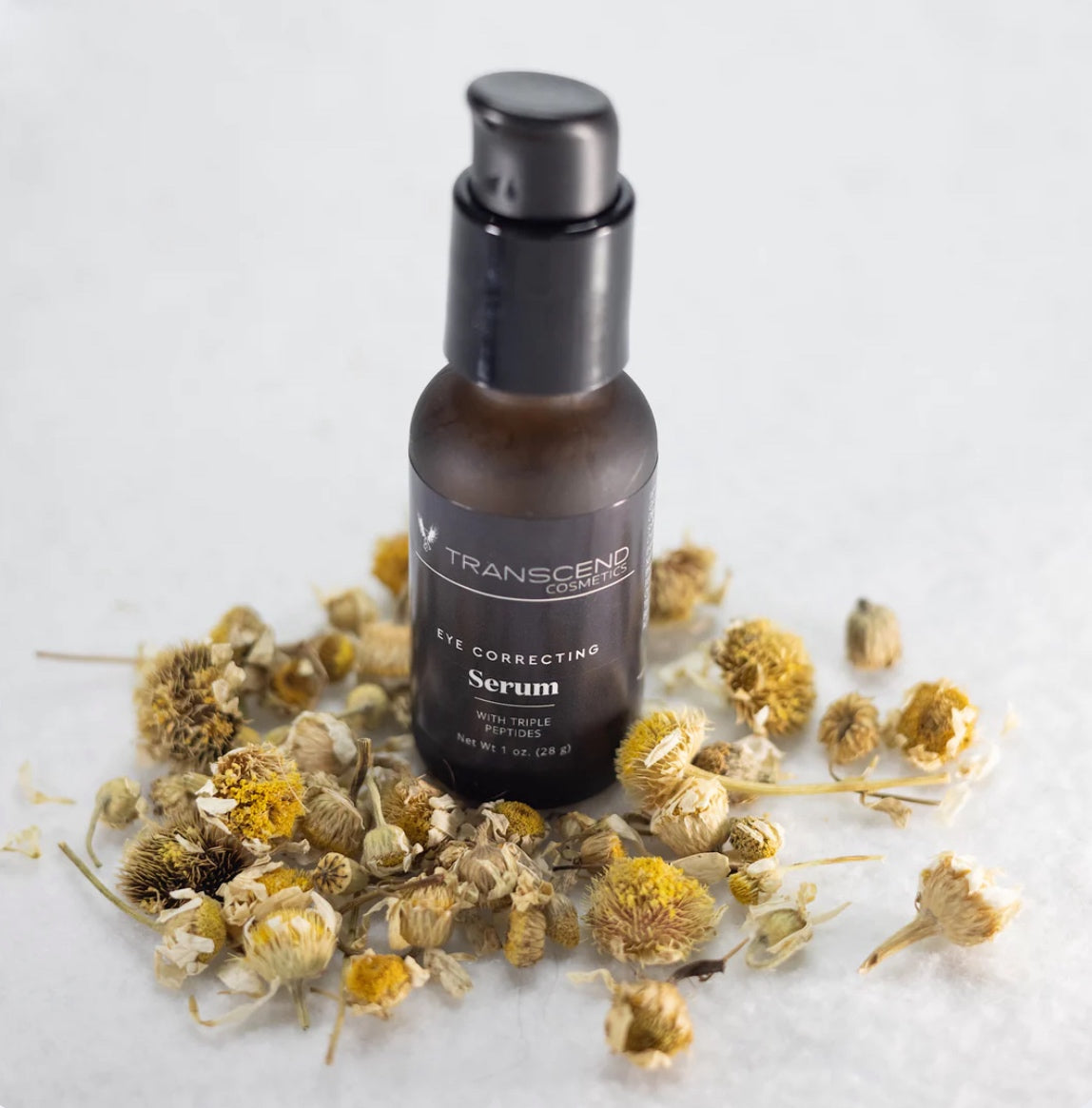 The image shows a dark amber bottle of Transcend Eye Correcting Serum, surrounded by dried chamomile flowers on a white background. The serum bottle features a black pump dispenser, and the label on the bottle indicates it contains chamomile and triple peptides. The flowers are scattered around the base of the bottle, emphasizing the natural ingredients theme.  Natural skincare made with chamomile. 
