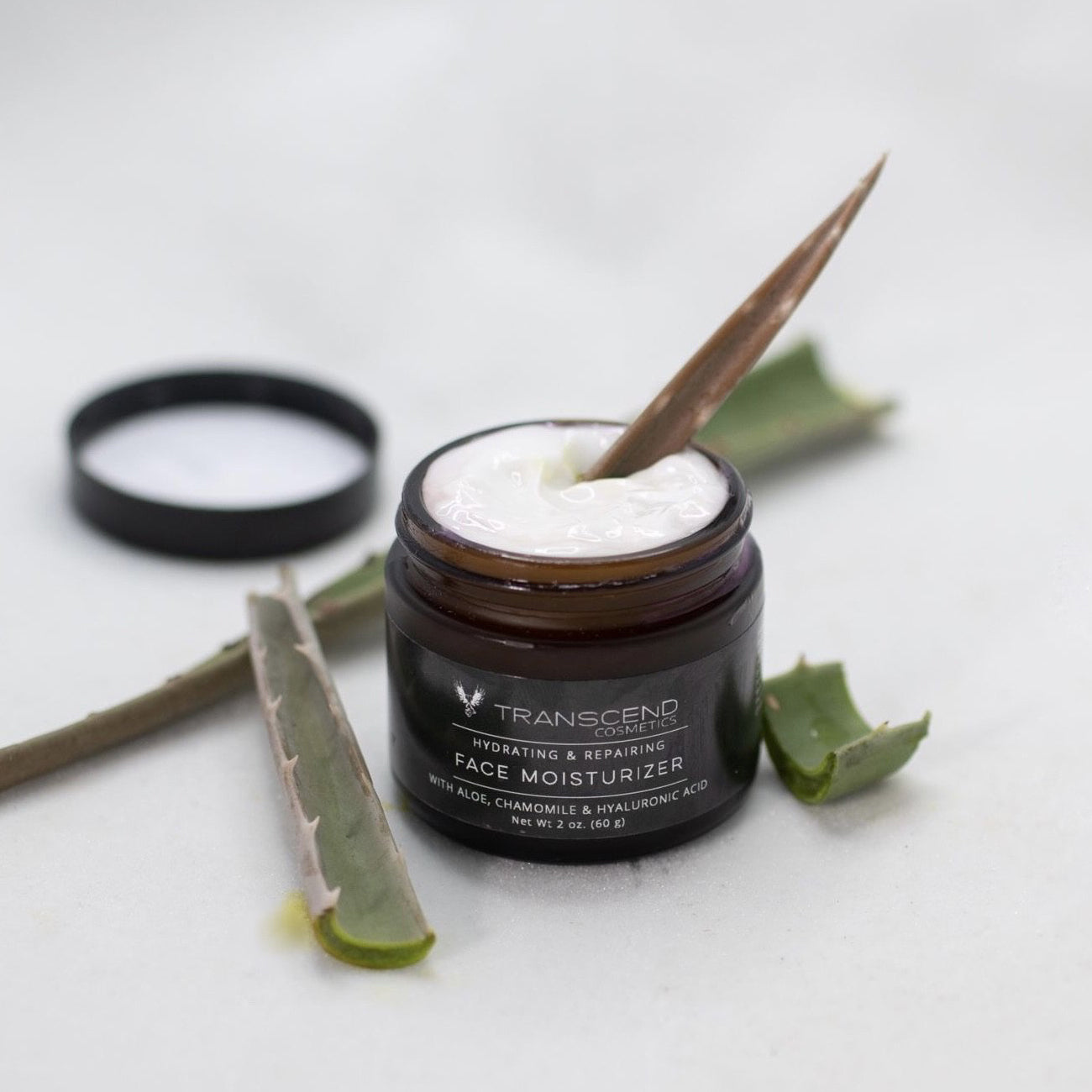 The image features a jar of Transcend Hydrating & Repairing Face Moisturizer on a light background. The moisturizer is creamy and white, with a aloe Vera leaf inside resting in the cream, lifting some of the product. Beside the jar, there are a few fresh aloe vera leaves, one of which is sliced open to show the gel inside. The product label indicates it contains aloe, chamomile, and hyaluronic acid. Natural skincare made with aloe Vera and chamomile 
