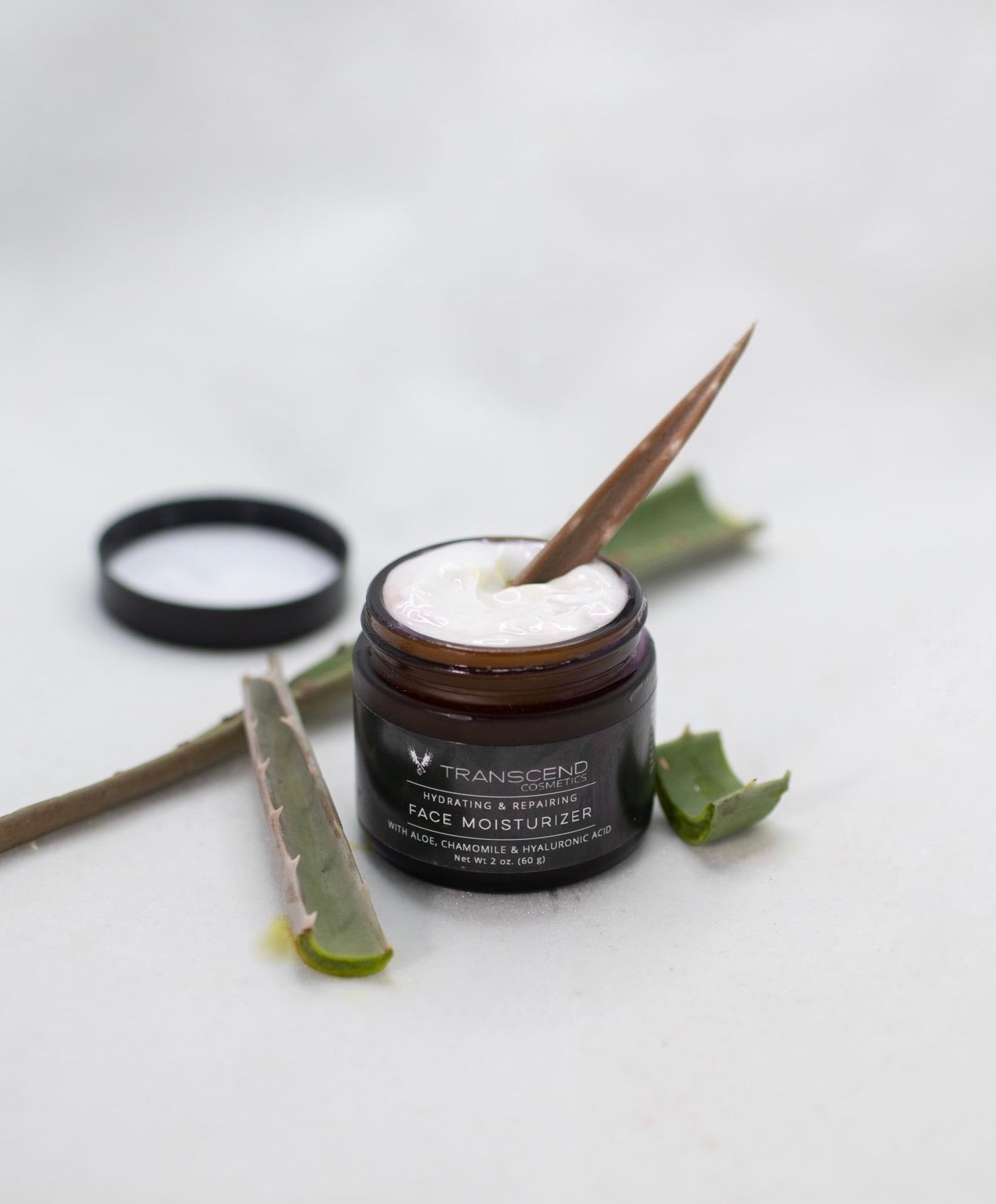 The image features a jar of Transcend Hydrating & Repairing Face Moisturizer on a light background. The moisturizer is creamy and white, with a aloe Vera leaf inside resting in the cream, lifting some of the product. Beside the jar, there are a few fresh aloe vera leaves, one of which is sliced open to show the gel inside. The product label indicates it contains aloe, chamomile, and hyaluronic acid.
