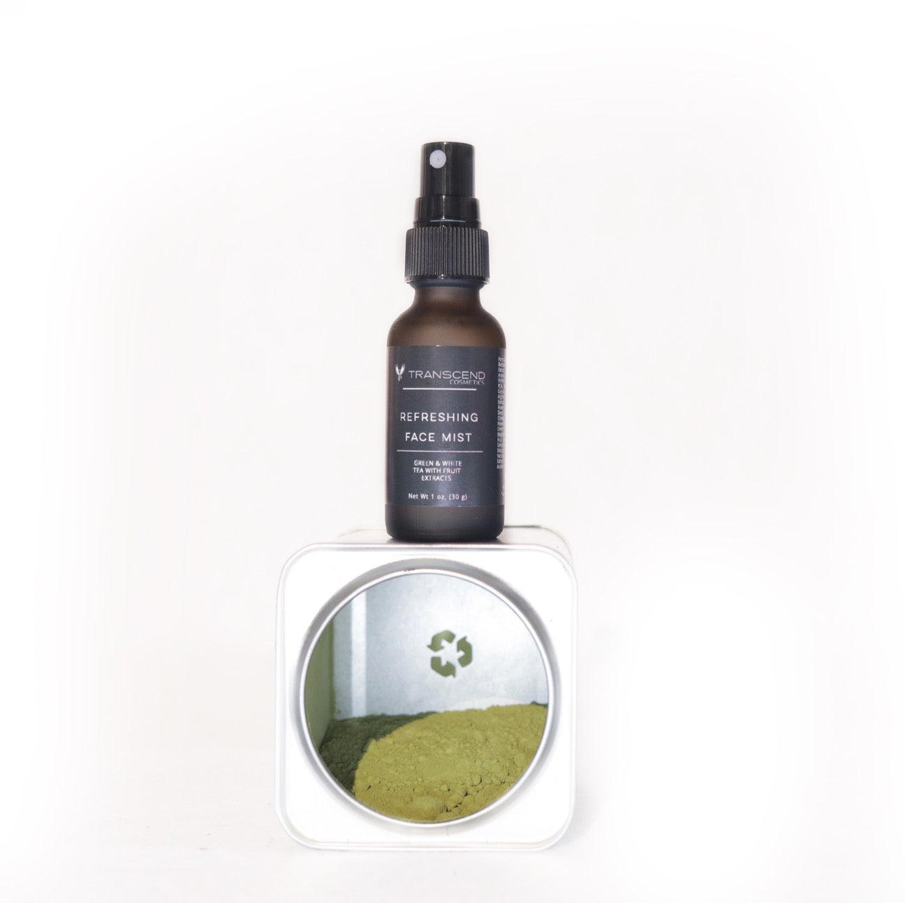 The image shows a bottle of Transcend Refreshing Face Mist on a light background. The face mist bottle is dark amber with a black spray nozzle and is labeled to indicate it contains green and white tea extracts. The bottle is placed on top of a container with an open lid, revealing green powder inside, highlighting the natural tea extract ingredient used in the face mist. Natural toner made with green tea 
