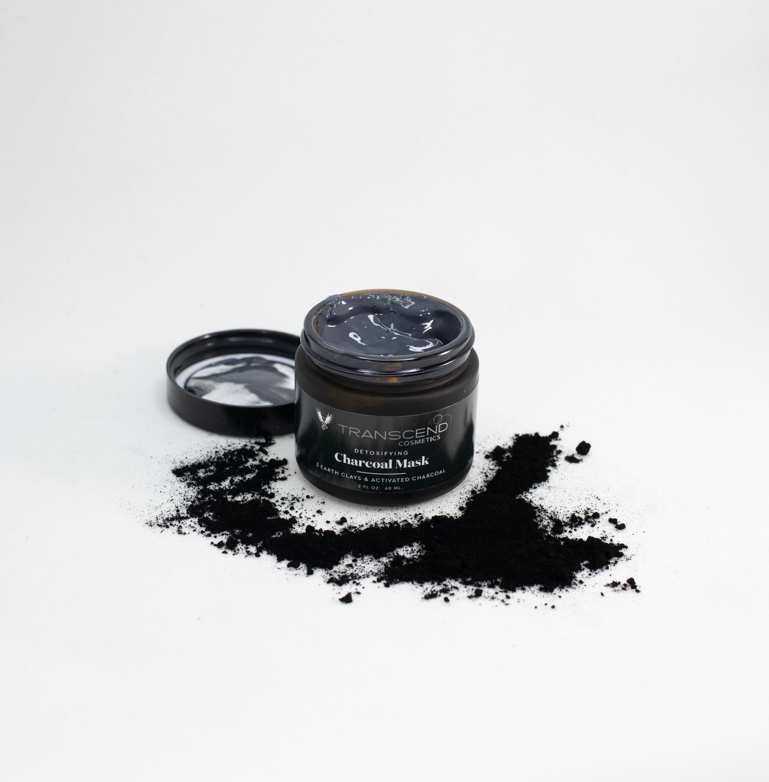 The image features a jar of Transcend Detoxifying Charcoal Mask open on a white background. The mask inside the jar is a dark, shiny black, contrasting with scattered black charcoal powder around the jar. The product's label clearly states that it contains kaolin clay and activated charcoal, emphasizing its purifying and detoxifying properties.
Natural skincare made with Kaolin Clay. 