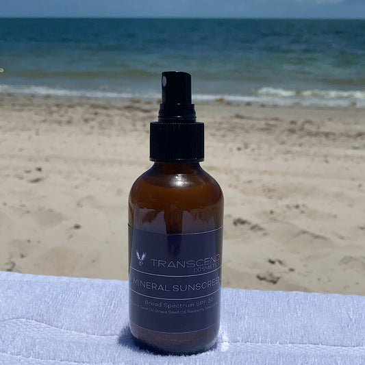 The image shows a bottle of Transcend Mineral Sunscreen placed on a towel at the beach, with the ocean and sandy shore in the background. The sunscreen bottle is dark amber with a black spray nozzle. The label indicates it provides broad-spectrum SPF 30 protection and contains natural ingredients like red raspberry seed oil, grape seed oil, zinc, and blueberry seed oil. The beach setting highlights the product's suitability for sun protection during outdoor activities.