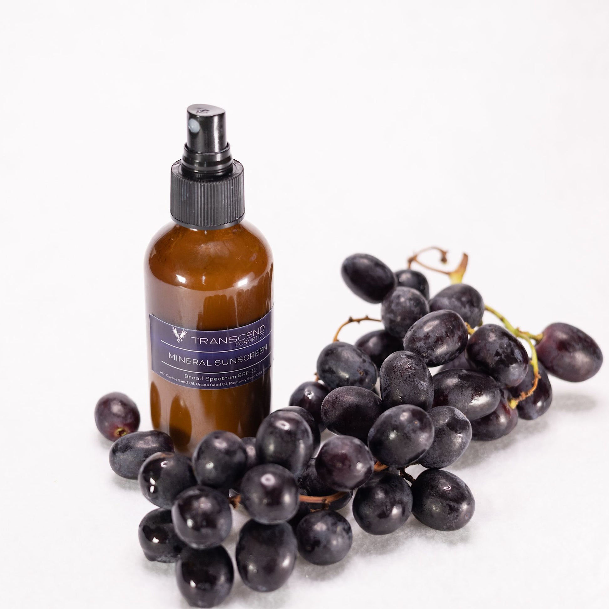 The image shows a bottle of Transcend Mineral Sunscreen on a light background. The sunscreen bottle is dark amber with a black spray nozzle. It is surrounded by a bunch of fresh black grapes, emphasizing the inclusion of grape seed oil in the product. The label on the bottle indicates it provides broad-spectrum SPF 30+ protection and contains natural ingredients like grape seed oil and blueberry extract. Organic Sunscreen made with Zinc 